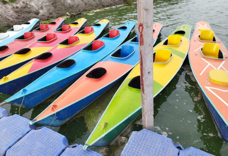 A row of colourful kayaks in red, yellow, blue, orange, and green rests alongside a dock on a greenish body of water. Th